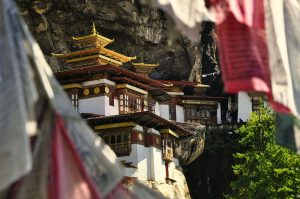 A close-up view of the Tiger's Nest Monastery (Paro Taktsang) in Bhutan, built into a dramatic cliffside, partially framed by colorful fluttering prayer flags in the foreground.